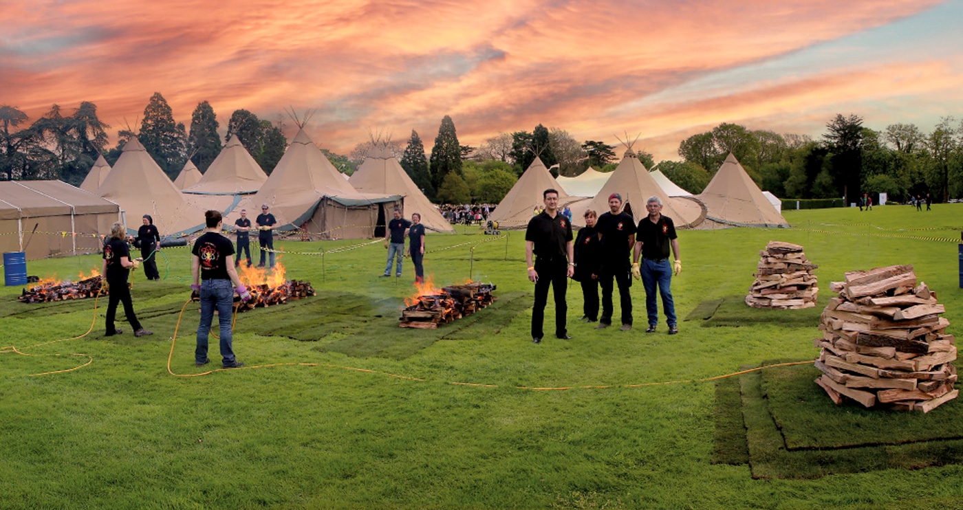 large group of firewalk team tending three fires standing in front of tents